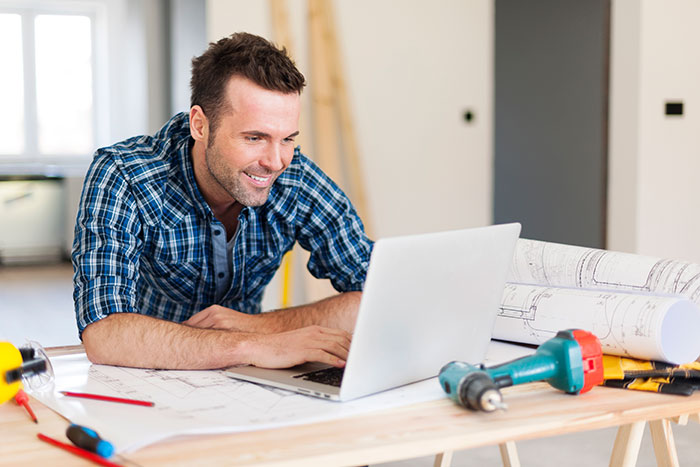 Man in plaid shirt working on laptop surrounded by tools and blueprints, representing financially trapped woman scenario. Man in plaid shirt working on laptop surrounded by tools and blueprints, representing financially trapped woman scenario.