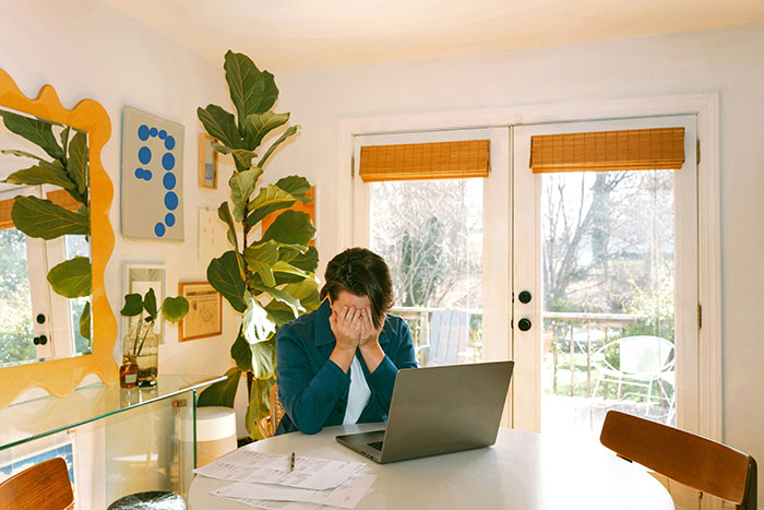 Woman feeling financially trapped sitting at table with laptop and paperwork in bright home, stressed and overwhelmed. Woman feeling financially trapped sitting at table with laptop and paperwork in bright home, stressed and overwhelmed.