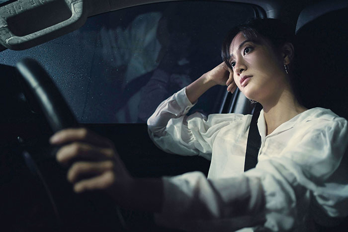 Woman sitting in a car at night, looking worried due to a flat tire causing her to miss work.
