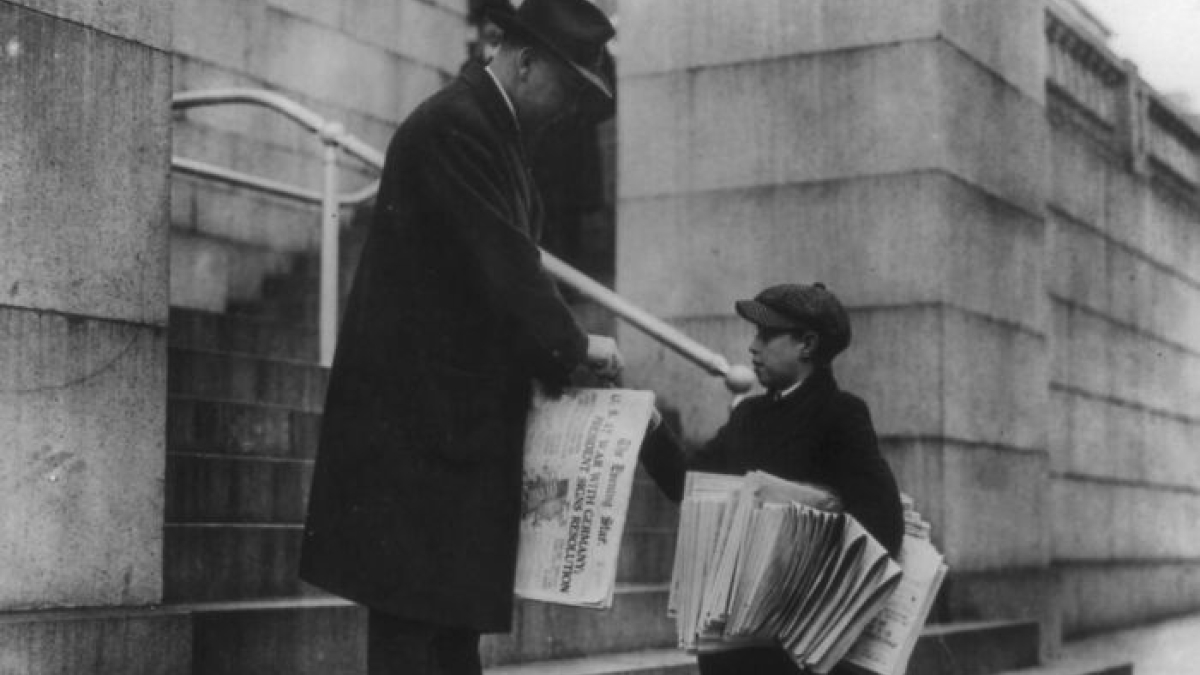 Boy selling newspapers on street to a man wearing a hat, illustrating daily life beyond the WWI trenches.