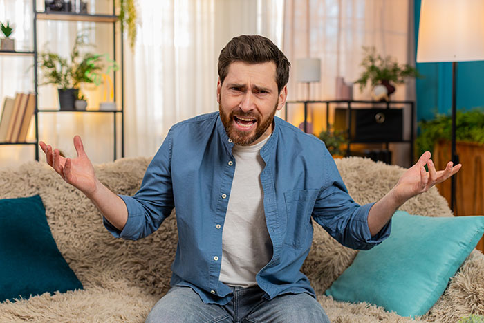 Man in blue shirt sitting on a couch, gesturing in frustration, illustrating teen escapes babysitting trap situation. Man in blue shirt sitting on a couch, gesturing in frustration, illustrating teen escapes babysitting trap situation.