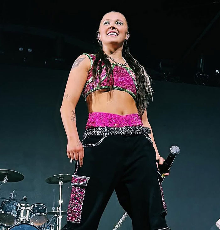 JoJo Siwa smiling on stage wearing a pink and black outfit during a live performance with microphone in hand. JoJo Siwa smiling on stage wearing a pink and black outfit during a live performance with microphone in hand.