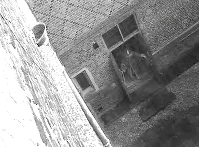 Black and white photo showing a tilted view of a brick building entrance with two figures, capturing strange and unexplained histories.