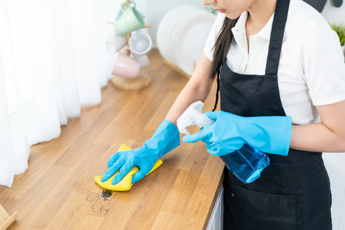 Woman wearing blue gloves and black apron cleaning a wooden countertop with a spray bottle and yellow cloth. Woman wearing blue gloves and black apron cleaning a wooden countertop with a spray bottle and yellow cloth.