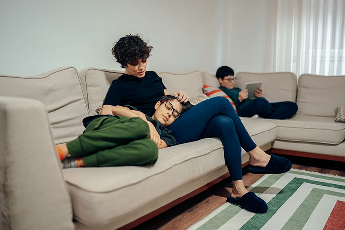 Woman comforting child on couch while another child uses tablet in living room, reflecting family tension at doorstep conflict. Woman comforting child on couch while another child uses tablet in living room, reflecting family tension at doorstep conflict.
