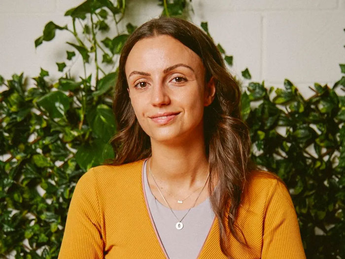 Woman with long brown hair wearing a mustard sweater, smiling calmly in front of green leafy plants indoors. Woman with long brown hair wearing a mustard sweater, smiling calmly in front of green leafy plants indoors.