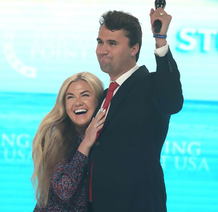 Man in suit holding microphone with woman smiling and hugging him at event, WWE level stuff controversy visible. Man in suit holding microphone with woman smiling and hugging him at event, WWE level stuff controversy visible.