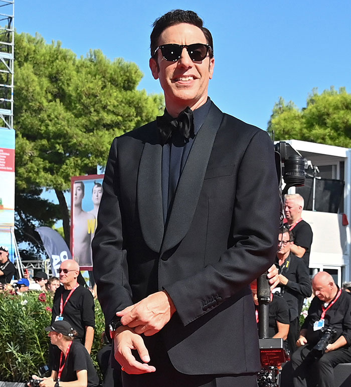 Sacha Baron Cohen wearing a black tuxedo and sunglasses at an outdoor event with people in the background Sacha Baron Cohen wearing a black tuxedo and sunglasses at an outdoor event with people in the background