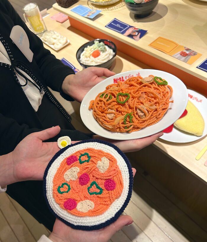 Crocheted realistic food accessories featuring pasta and pizza held by two people, showcasing Japanese fiber art.