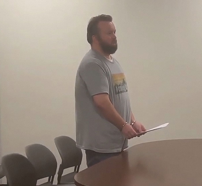 Man in handcuffs stands by a table in a plain room during a teacher arrest related to poop spray incident at school. Man in handcuffs stands by a table in a plain room during a teacher arrest related to poop spray incident at school.