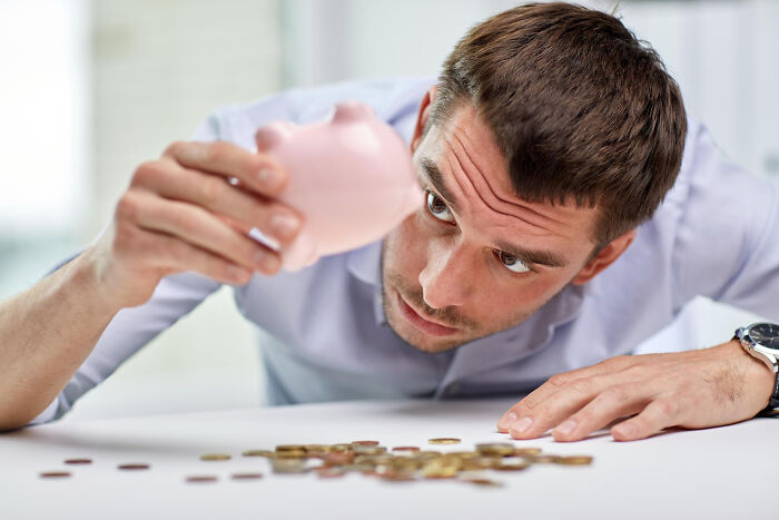 Man inspecting a nearly empty piggy bank with scattered coins, reflecting on mind-boggling conversations about adult life.