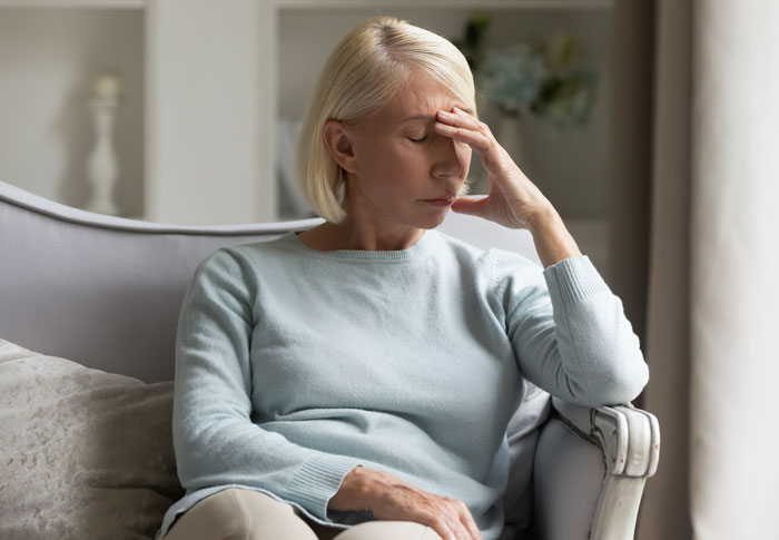 Middle-aged woman in a sweater sitting on a chair, looking distressed and holding her forehead in a living room. Middle-aged woman in a sweater sitting on a chair, looking distressed and holding her forehead in a living room.