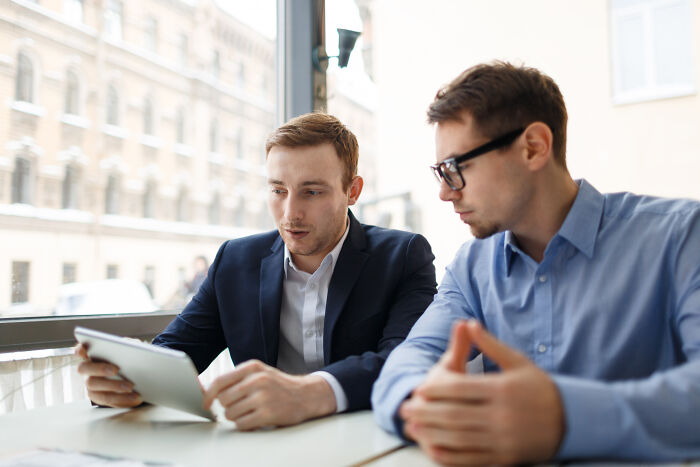 Two young men in business attire having a mind-boggling conversation while looking at a digital tablet in a bright office.