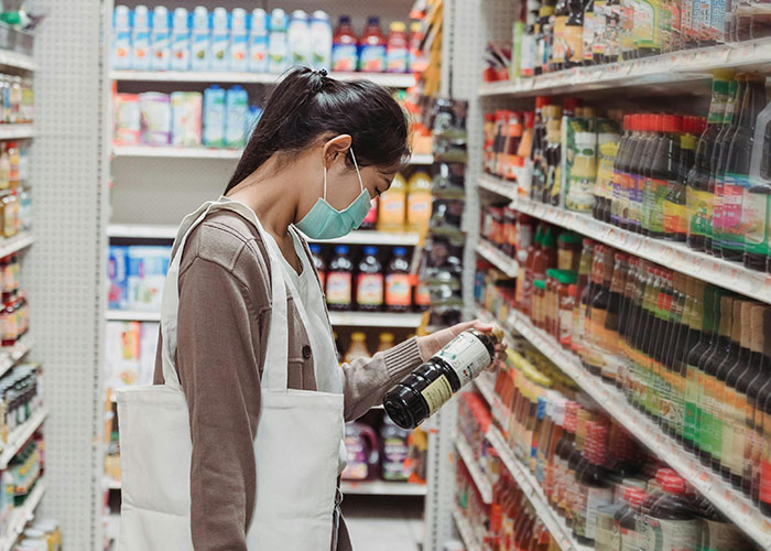 Woman with a tote bag wearing a face mask, examining products on a grocery store shelf, reflecting common purchases and pricing concerns.