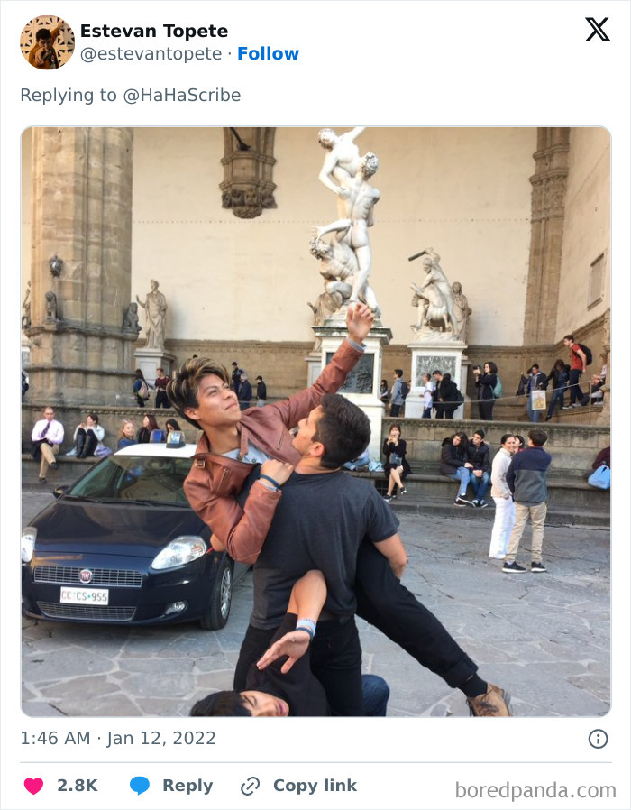 Two men creatively imitating an iconic art statue pose outdoors with a crowd and historic sculptures in the background.