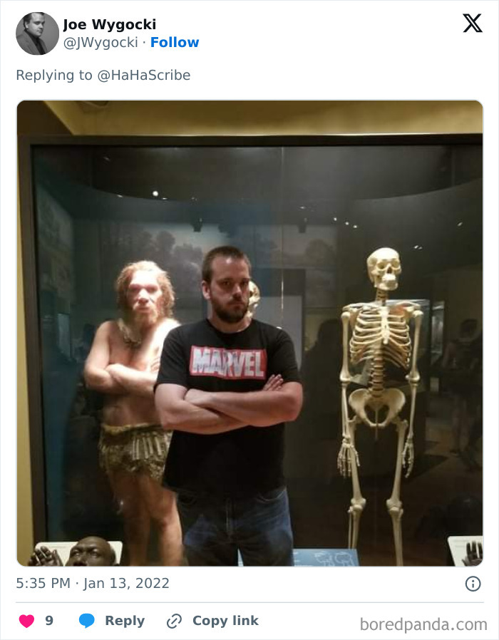 Man in Marvel shirt imitating prehistoric figure pose next to skeleton and ancient human display in a museum setting.