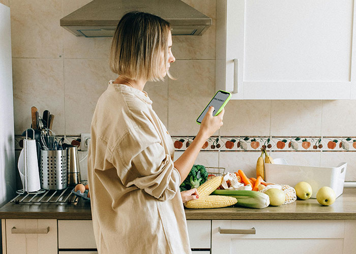 Woman in kitchen checking phone while surrounded by fresh vegetables, illustrating common purchases ditched due to price tags