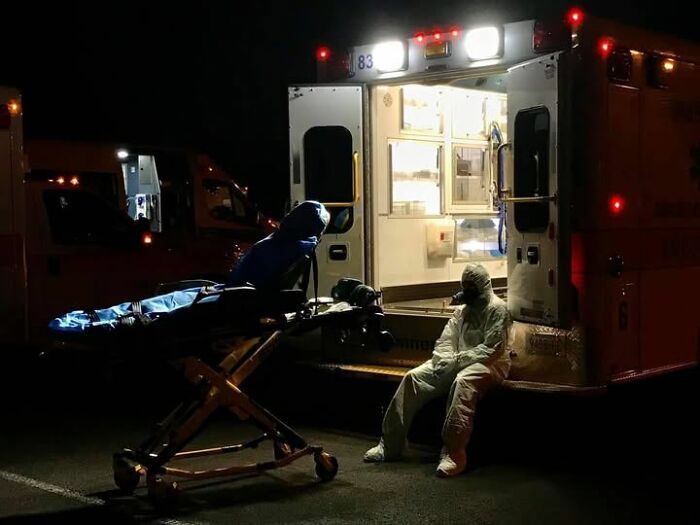Paramedic in protective gear resting by an open ambulance at night with a stretcher nearby, EMT scene after a call.