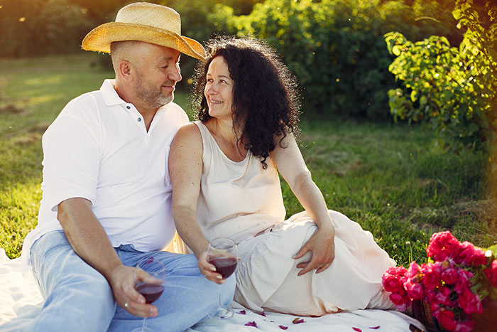 Couple sitting on a picnic blanket outdoors enjoying wine, with sunlight and greenery in the background on a calm day. Couple sitting on a picnic blanket outdoors enjoying wine, with sunlight and greenery in the background on a calm day.