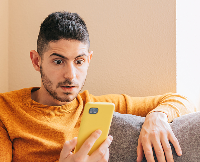 Man wearing orange sweater looking shocked at phone while sitting on a couch, depicting relationship trust issues. Man wearing orange sweater looking shocked at phone while sitting on a couch, depicting relationship trust issues.