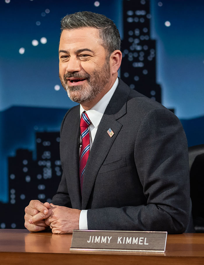 Jimmy Kimmel in a suit and tie, seated at a desk during a television show segment about his show suspension. Jimmy Kimmel in a suit and tie, seated at a desk during a television show segment about his show suspension.