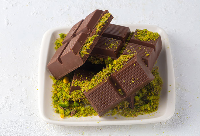 Dark chocolate pieces topped with bright green crushed pistachios served in a white square dish.