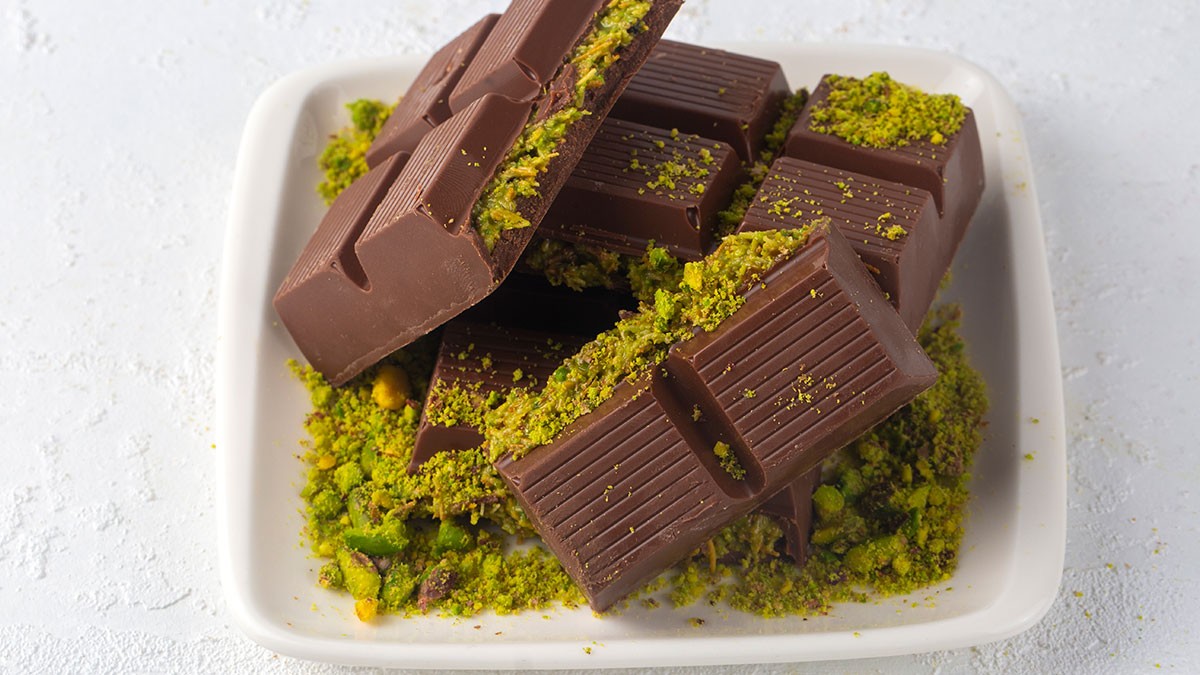 Chocolate bars topped with crushed pistachios arranged on a white square plate with a light textured background.