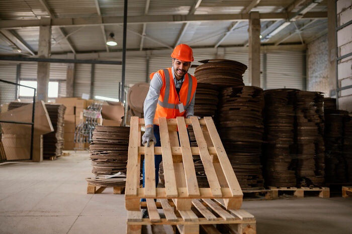 Warehouse worker in safety gear handling wooden pallets amid stacks of materials, reflecting mind-boggling adult conversations.