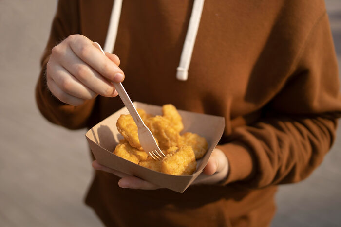 Person in brown hoodie holding a tray of chicken nuggets using a wooden fork at an outdoor school incident location