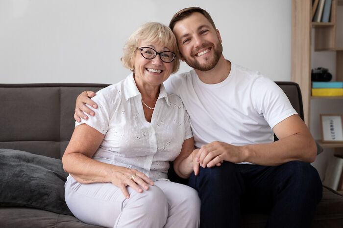 Elderly mother-in-law smiling with adult son on couch, illustrating entitled lady annoyed as MIL splits estate half to hubby and niece.