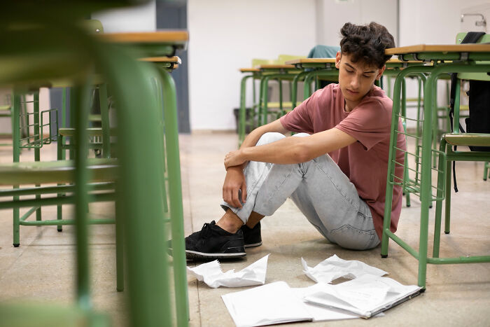 Teenage boy sitting on classroom floor looking upset with scattered school papers, representing shocking school incidents.