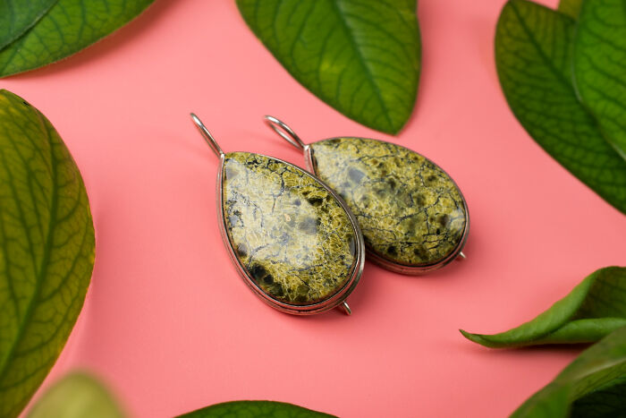 Pair of green gemstone earrings on a pink background surrounded by green leaves, symbolizing hexes and witchcraft charms.
