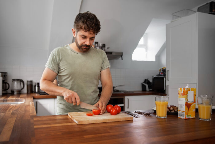 Man cutting tomatoes in kitchen, reflecting on moments of falling out of love, surrounded by orange juice and utensils.
