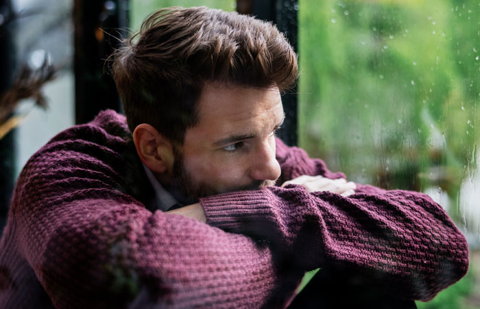 Young man in a maroon sweater looking out a window, reflecting on family expectations and caring for autistic brother. Young man in a maroon sweater looking out a window, reflecting on family expectations and caring for autistic brother.