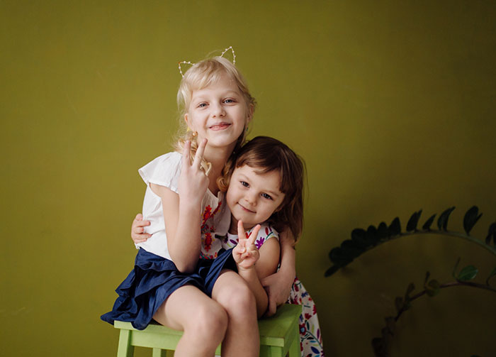 Two young girls sitting together on a green stool, smiling and making peace signs against an olive backdrop.