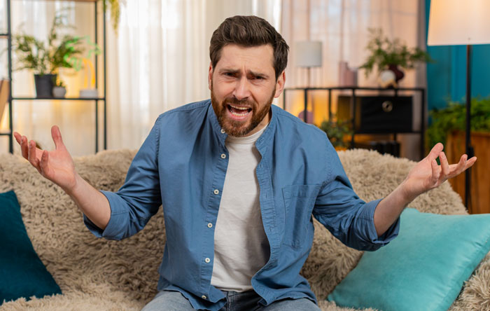 Frustrated man sitting on couch with arms open, expressing regret and tension in a cozy living room setting. Frustrated man sitting on couch with arms open, expressing regret and tension in a cozy living room setting.