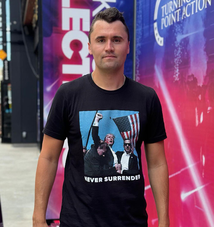 Charlie Kirk wearing a black Never Surrender t-shirt standing outside with Turning Point Action signage in the background. Charlie Kirk wearing a black Never Surrender t-shirt standing outside with Turning Point Action signage in the background.