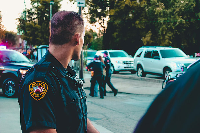 Police officer in uniform observing a tense street scene involving teens and multiple police vehicles at dusk. Police officer in uniform observing a tense street scene involving teens and multiple police vehicles at dusk.