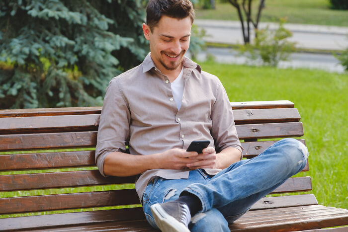 Young man sitting on a park bench smiling while using a smartphone, illustrating childish reasons ladies ditched a guy.