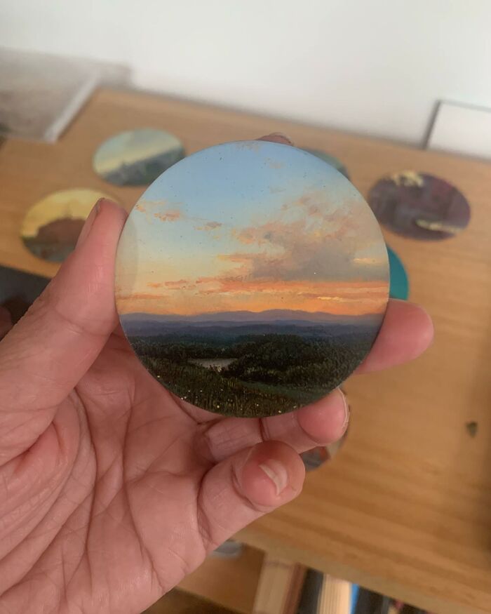 Hand holding a detailed painting by Dina Brodsky of a natural landscape with a sunset and distant mountains.