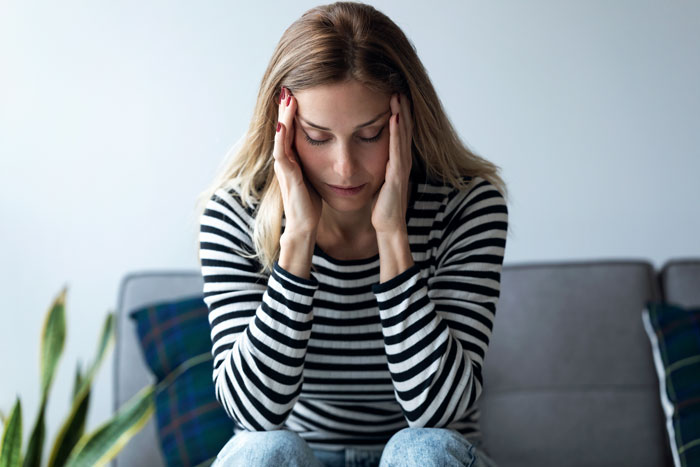 Stressed single sister in striped shirt sitting on couch struggling to handle sibling's engagement news in living room. Stressed single sister in striped shirt sitting on couch struggling to handle sibling's engagement news in living room.