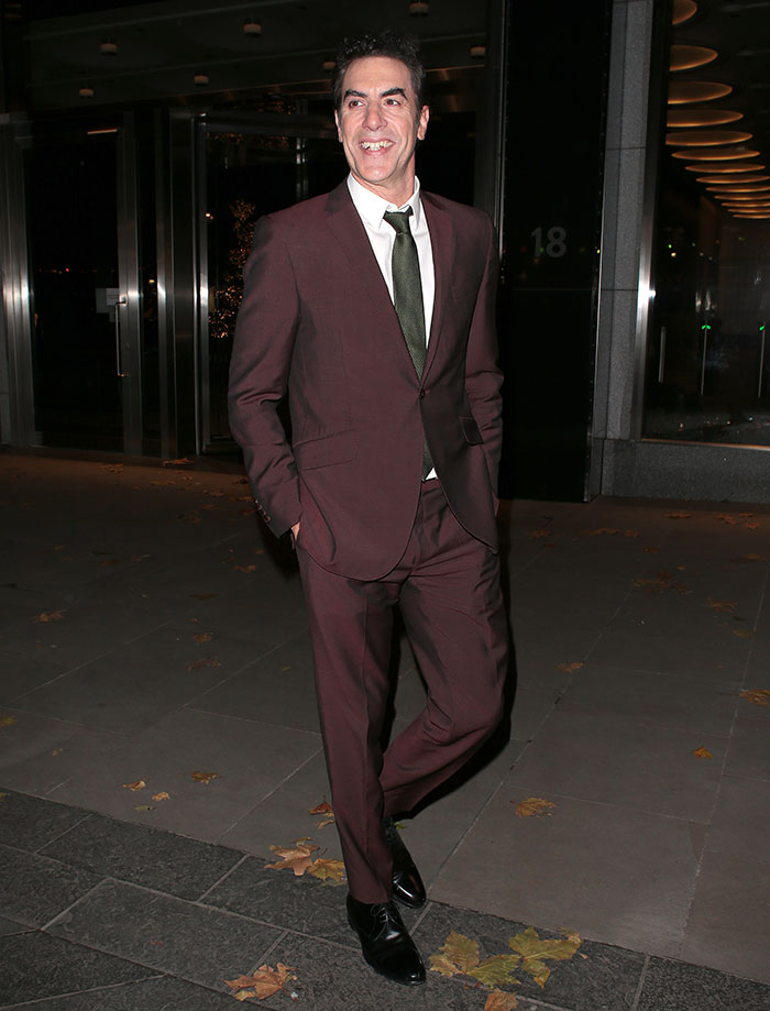 Sacha Baron Cohen, 53, in a burgundy suit walking outside at night after $160M divorce from Isla Fisher. Sacha Baron Cohen, 53, in a burgundy suit walking outside at night after $160M divorce from Isla Fisher.