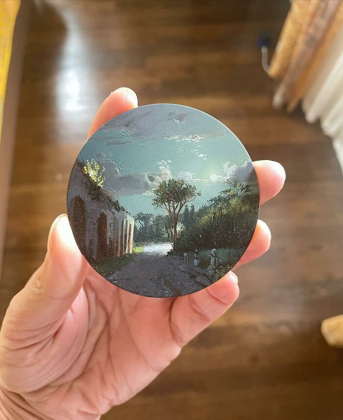 Hand holding a detailed painting by Dina Brodsky depicting a natural landscape with trees and a cloudy sky.