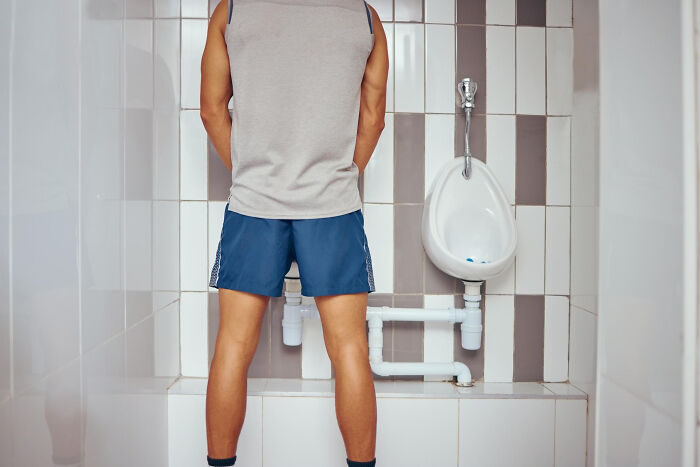 Man standing in a public restroom wearing shorts and a tank top, facing a urinal against a tiled wall.
