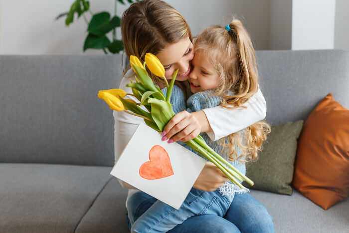 Woman and child hugging on a couch, holding yellow tulips and a heart card, symbolizing love and relationships.