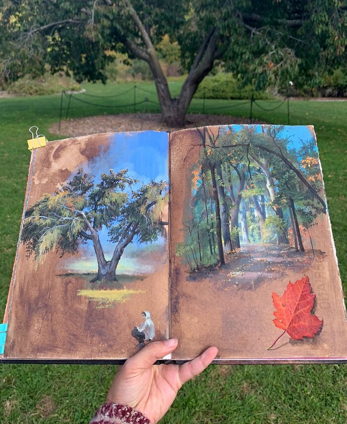 Hand holding open sketchbook with detailed paintings by Dina Brodsky of trees and a forest path showing natural world beauty.