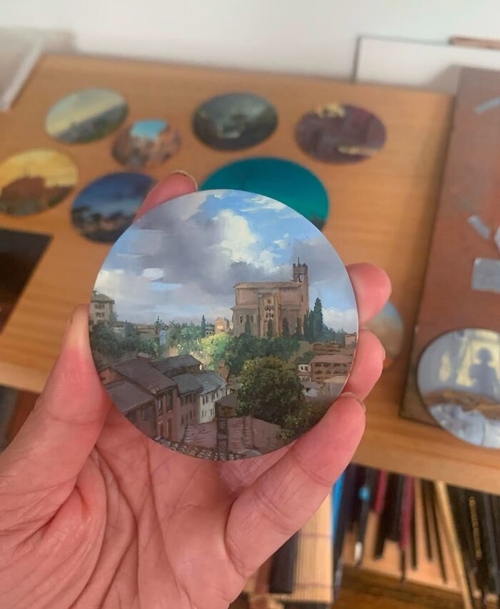 Hand holding a detailed circular painting by Dina Brodsky depicting a natural landscape with buildings and cloudy sky.