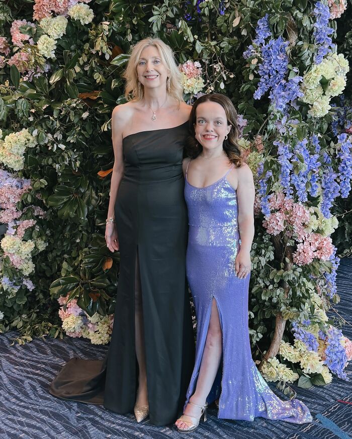 31YO woman with dwarfism in a sparkling purple gown posing with a taller woman in a black dress against a floral backdrop 31YO woman with dwarfism in a sparkling purple gown posing with a taller woman in a black dress against a floral backdrop