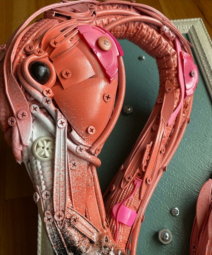 Pink flamingo animal sculpture made from repurposed trash items, displayed in an ornate white frame against a wooden background.