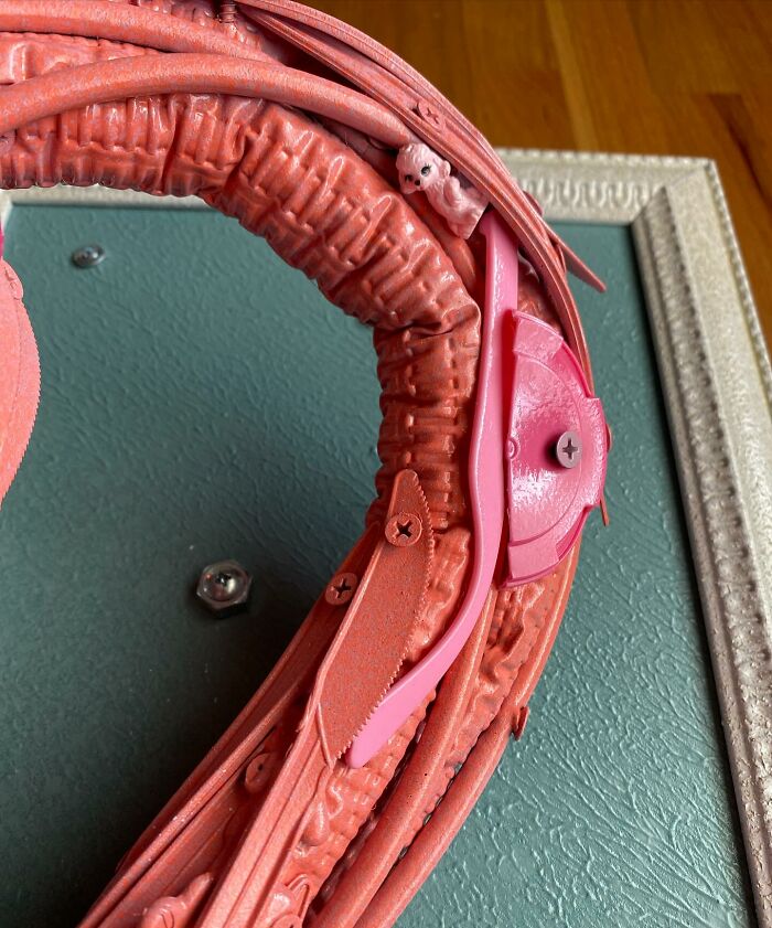 Pink flamingo animal sculpture made from repurposed trash items, displayed in an ornate white frame against a wooden background.